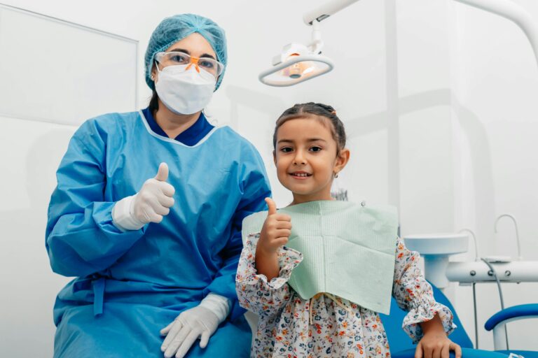 Smiling child giving a thumbs-up in a dental clinic beside a friendly dentist, with a dental chair visible in the background.