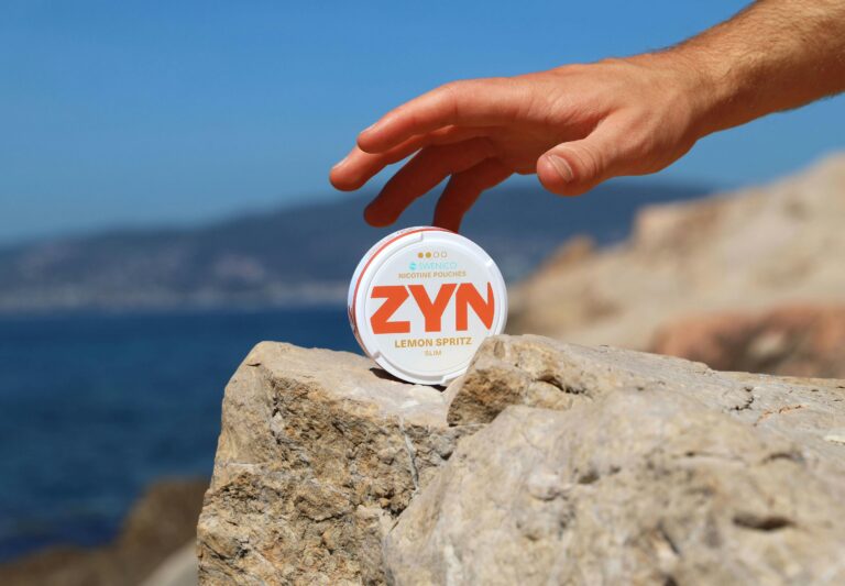 A hand reaching for a Zyn nicotine pouch box placed on a mountain rock with a blue ocean in the background.